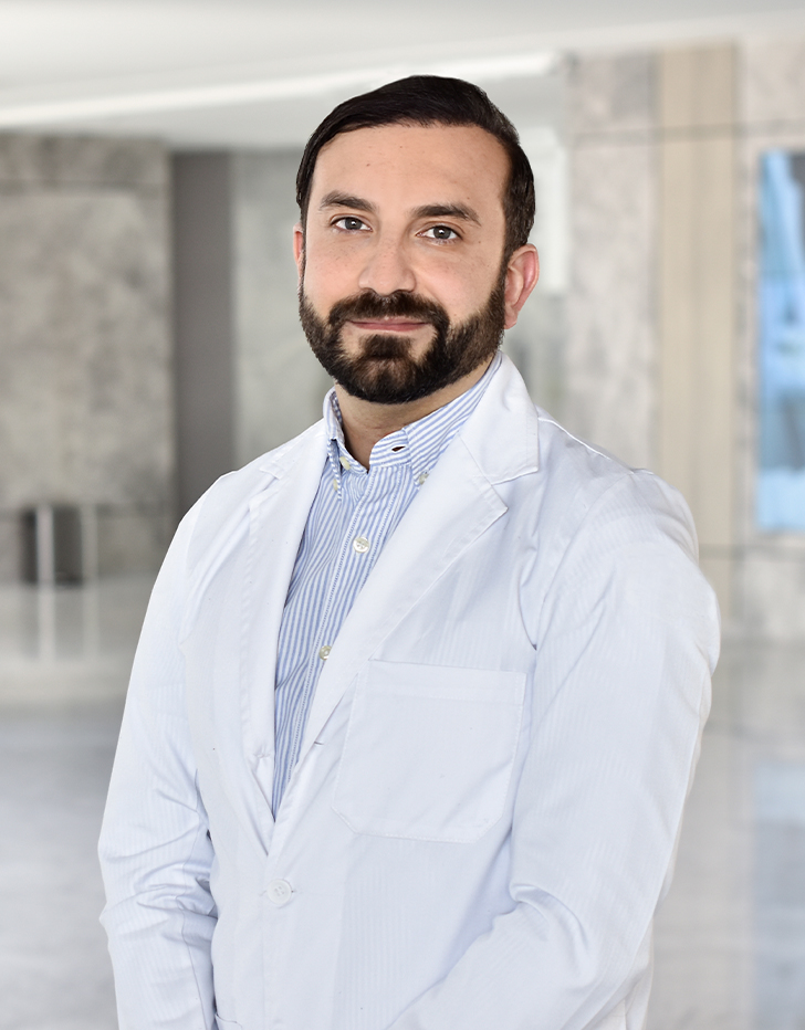 Es la foto de perfil de un oncólogo médico en Querétaro de Hospital Angeles Centro Sur. Usa una bata y una camisa de rayas color azul, tiene barba y de fondo es la recepción del hospital.