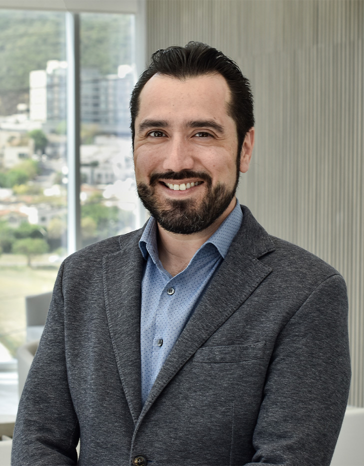 Es la foto de perfil de un pediatra y neonatólogo en San Pedro Garza de Hospital Angeles Valle Oriente.  Usa un saco gris con una camisa azul. Tiene el cabello y la barba negra. Está sonriendo y sentado.