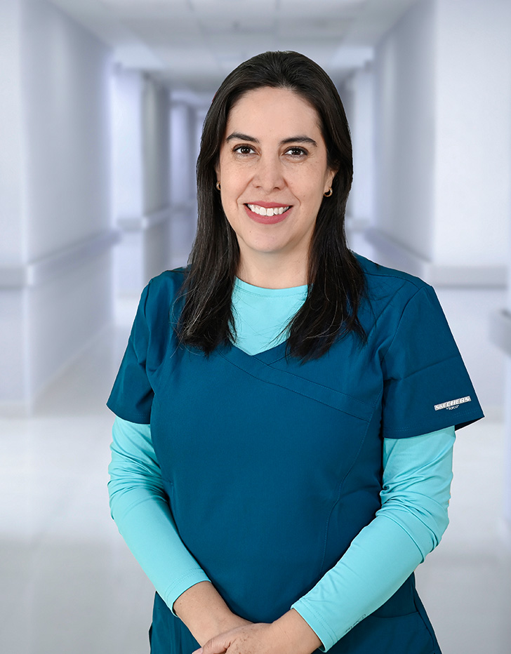 Es la foto de perfil de un odontólogo en Ciudad Juárez de Hospital Angeles Ciudad Juárez. La doctora tiene el cabello oscuro y lacio, y está sonriendo. Lleva una bata médica blanca sobre una blusa negra. En sus orejas, se pueden ver pequeños aretes de perla. El fondo muestra un área luminosa del hospital, con paredes claras y una pintura en tonos cálidos.