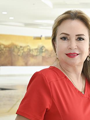 Es la foto de perfil de un especialista en audiología y foniatría en Ciudad Juárez de Hospital Angeles Ciudad Juárez. Trae un uniforme rojo y tiene el cabello largo y rubio. Utiliza una bata blanca, una blusa azul y tiene cabello negro.