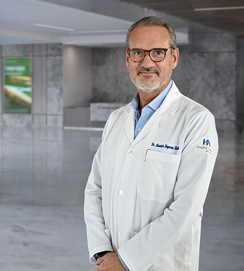 Es la foto de perfil de un ortopedista y traumatólogo en Querétaro de Hospital Angeles Centro Sur. El doctor tiene una bata blanca y una camisa gris tiene barba canosa y lentes con marco negro.