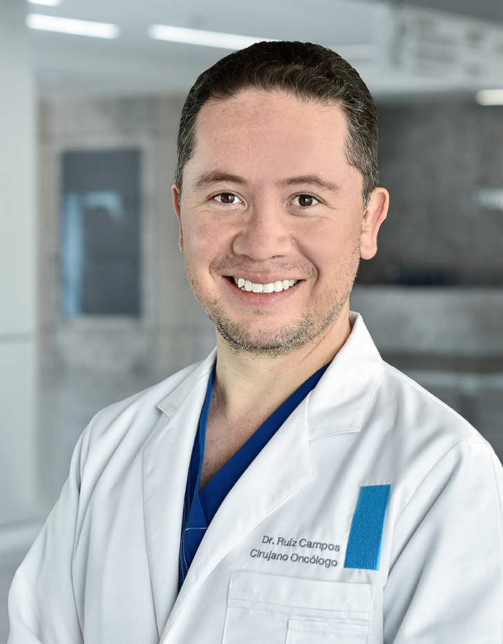 Es la foto de perfil de un cirujano oncológico en Querétaro de Hospital Angeles Centro Sur. Viste una bata blanca y una camisa azul está sonriendo.