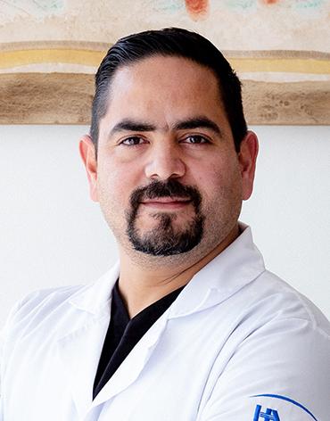 Es la foto de perfil de un ginecólogo y obstetra en Ciudad Juárez de Hospital Angeles Ciudad Juárez. El doctor es un hombre con barba y bigote, cabello oscuro peinado hacia atrás. Está vestido con una bata médica blanca y debajo lleva una camiseta negra. En la manga de la bata se distingue un logotipo de un hospital. El fondo es claro, con una pintura en tonos suaves en la parte superior.