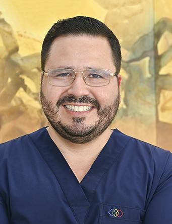 Es la foto de perfil de un radiólogo en Ciudad Juárez de Hospital Angeles Ciudad Juárez. El doctor tiene barba y bigote, cabello oscuro y corto, y lleva gafas. Está sonriendo y viste un uniforme médico de color azul marino con un pequeño logotipo del hospital en el pecho. El fondo muestra una pintura en tonos cálidos con figuras abstractas, creando un ambiente acogedor.