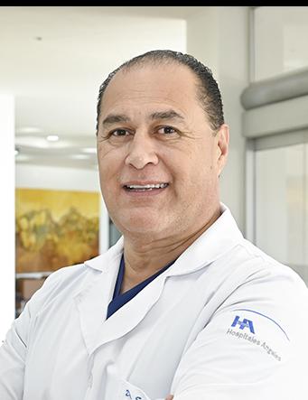 Es la foto de perfil de un odontólogo en Ciudad Juárez de Hospital Angeles Ciudad Juárez. El doctor tiene el cabello corto y oscuro, con una expresión amigable mientras sonríe. Lleva una bata médica blanca con el logotipo del Hospital Ángeles en la manga. Debajo de la bata, viste una camiseta azul. El fondo muestra un área iluminada del hospital con paredes claras y una pintura en tonos cálidos.