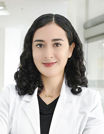 Es la foto de perfil de un psiquiatra en Ciudad Juárez de Hospital Angeles Ciudad Juárez. La doctora tiene el cabello oscuro, rizado y suelto. Está sonriendo y viste una bata médica blanca sobre una camiseta negra. Lleva un collar dorado con un nombre grabado. El fondo muestra un área luminosa del hospital, con paredes claras y un diseño moderno.