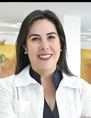 Es la foto de perfil de un odontólogo en Ciudad Juárez de Hospital Angeles Ciudad Juárez. La doctora tiene el cabello oscuro y lacio, y está sonriendo. Lleva una bata médica blanca sobre una blusa negra. En sus orejas, se pueden ver pequeños aretes de perla. El fondo muestra un área luminosa del hospital, con paredes claras y una pintura en tonos cálidos.