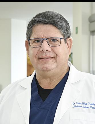 Es la foto de perfil de un pediatra en Ciudad Juárez de Hospital Angeles Ciudad Juárez. El doctor tiene el cabello canoso, corto, y lleva gafas. Está sonriendo levemente y viste una bata médica blanca sobre un uniforme azul marino. Su nombre y la especialidad 'Medicina Interna' están bordados en el pecho de la bata. El fondo muestra un área luminosa del hospital con paredes claras y una pintura en tonos cálidos.