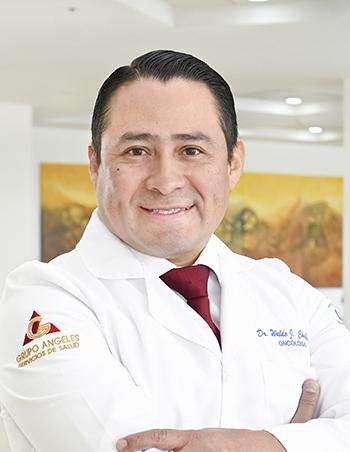 Es la foto de perfil de un cirujano oncológico en Ciudad Juárez de Hospital Angeles Ciudad Juárez. El doctor tiene el cabello oscuro y corto, peinado hacia atrás. Está sonriendo y lleva una bata médica blanca con su nombre y la especialidad 'Oncología Ginecológica' bordados en el pecho, junto con una corbata roja. En la manga de la bata se distingue el logotipo de 'Grupo Ángeles Servicios de Salud'. El fondo muestra un área luminosa del hospital, con paredes claras y una pintura en tonos cálidos.