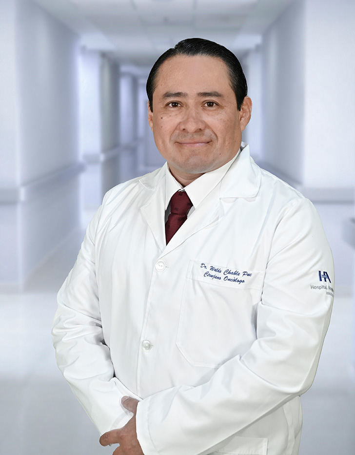 Es la foto de perfil de un cirujano oncológico en Ciudad Juárez de Hospital Angeles Ciudad Juárez. El doctor tiene el cabello oscuro y corto, peinado hacia atrás. Está sonriendo y lleva una bata médica blanca con su nombre y la especialidad 'Oncología Ginecológica' bordados en el pecho, junto con una corbata roja. En la manga de la bata se distingue el logotipo de 'Grupo Ángeles Servicios de Salud'. El fondo muestra un área luminosa del hospital, con paredes claras y una pintura en tonos cálidos.
