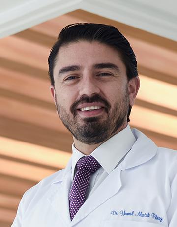 Es la foto de perfil de un neurólogo en Querétaro de Hospital Angeles Centro Sur. Usa una bata blanca, con una camisa blanca y una corbata morada con lunares blancos. Tiene barba y está sonriendo.