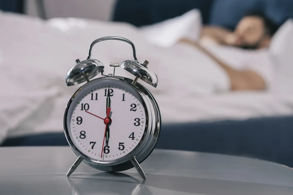 Reloj despertador en primer plano y hombre en cama que padece trastornos del sueño atendidos en la Clínica del Sueño en Hospital Angeles Lomas. 

