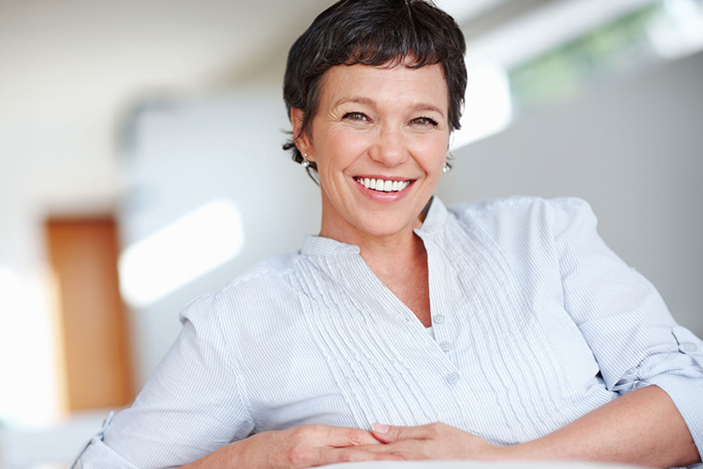 Mujer de 40 años, cabello negro, tez clara; cruzada de brazos y sonriendo dentro de cuarto difuminado color blanco.