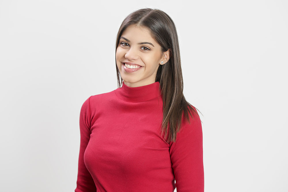 Mujer joven con sueter rojo sonriendo y viendo fijamente hacia enfrente en fondo color beige.