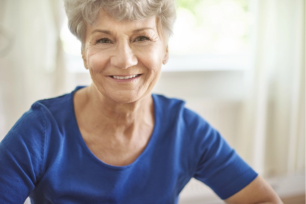 Mujer de la tercera edad, cabello blanco y blusa azul marino, mirando y sonriendo hacia enfrente sobre fondo difuminado color blanco con amarillo.