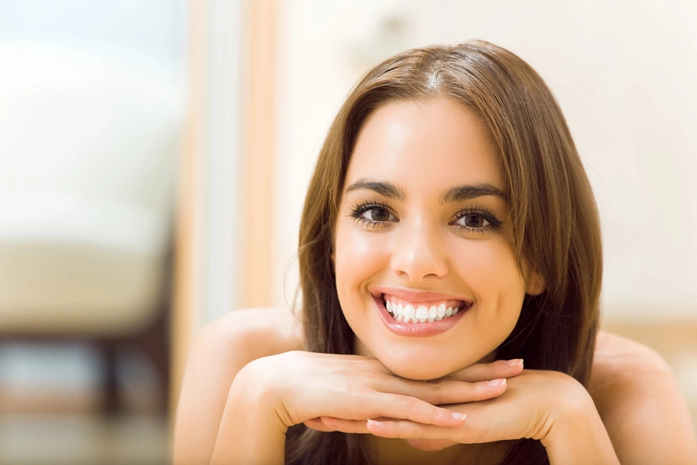 Mujer joven de cabello castaño, tez clara sonriendo y viendo fijamente hacia enfrente mientras apoya su rostro sobre sus dos manos.