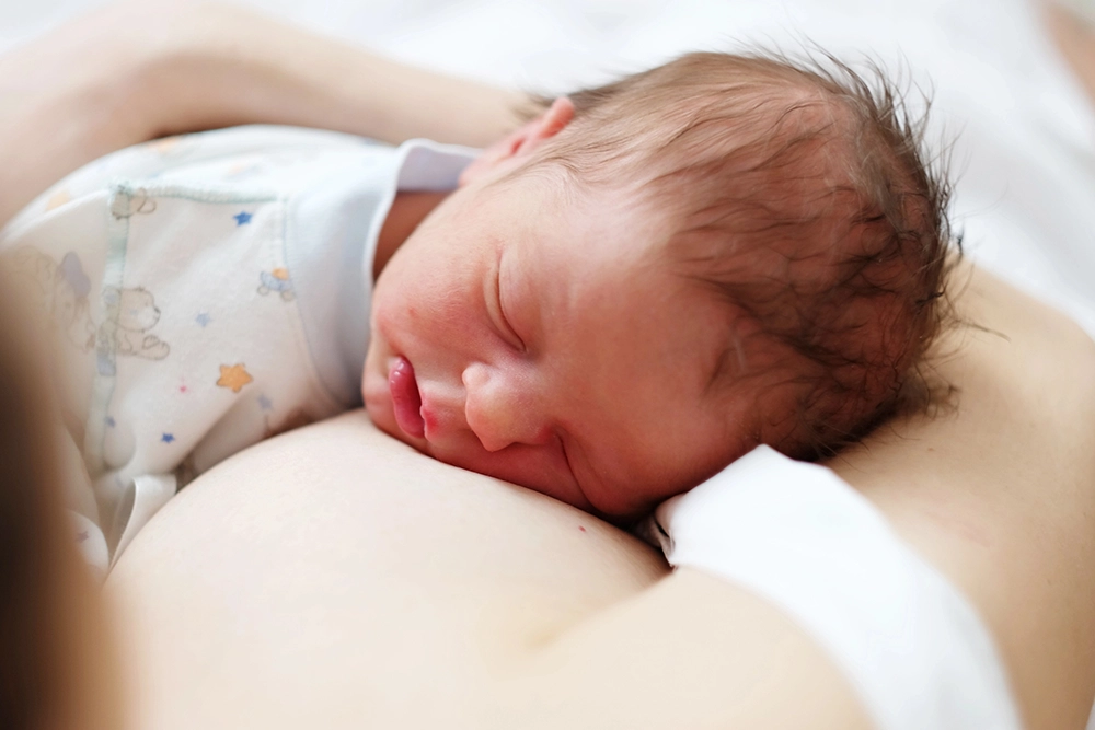 Bebé de tez clara, cabello café con mameluco blanco, dormido sobre el pecho de su mamá.