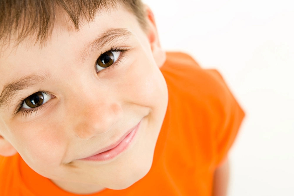 Primer plano de rostro de niño de cabello café, playera anaranjada viendo fijamente y sonriendo hacia arriba.