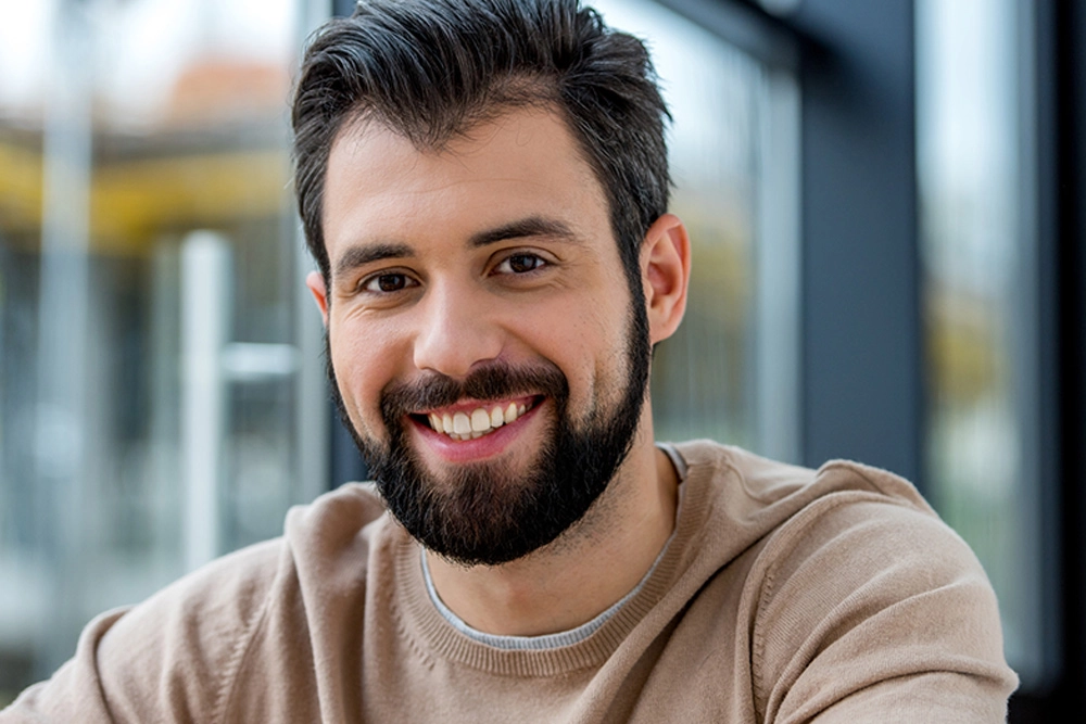 Hombre joven de cabello negro,barba negra y sueter café sonriendo en un fondo difuminado de color gris con amarillo.