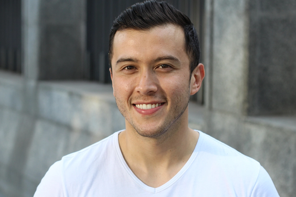 Hombre joven de cabello negro, barba, playera blanca con una sonrisa viendo fijamente hacia enfrente en fondo gris.