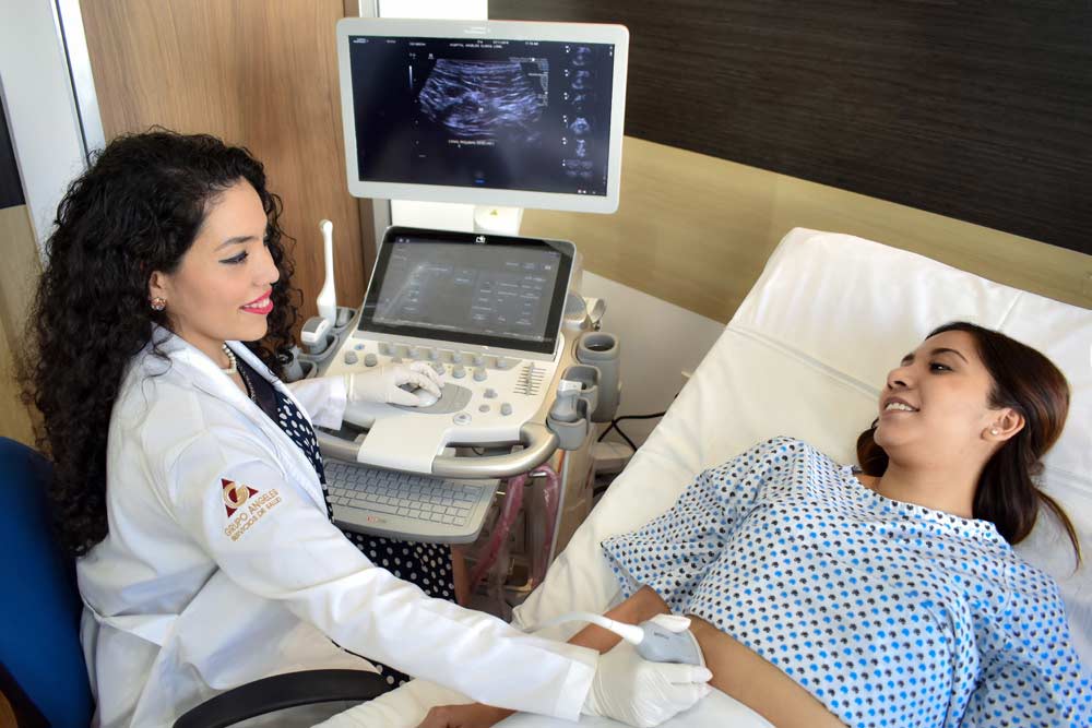 Doctora explorando con el trasductor el vientre de una paciente embarazada recostada en la cama de exploración.
