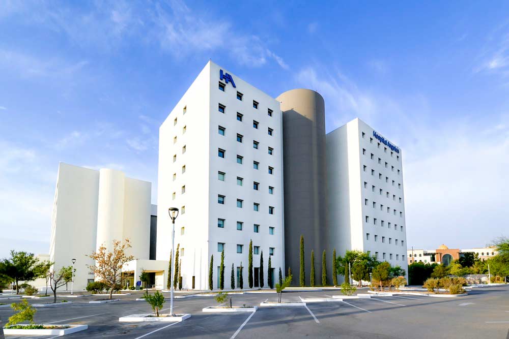 Fachada exterior con torres blancas y una en color gris del Hospital Angeles Ciudad Juárez y logotipo de HA en color azul.